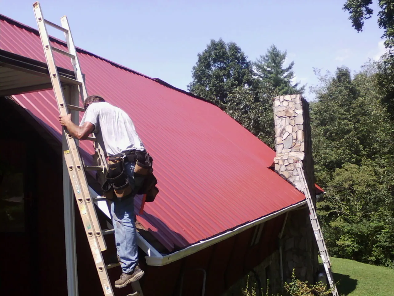 Roofer on a ladder beside a red metal roof and stone chimney — Soffit & Fascia Repair by Roofing Brothers in Snow Shoe